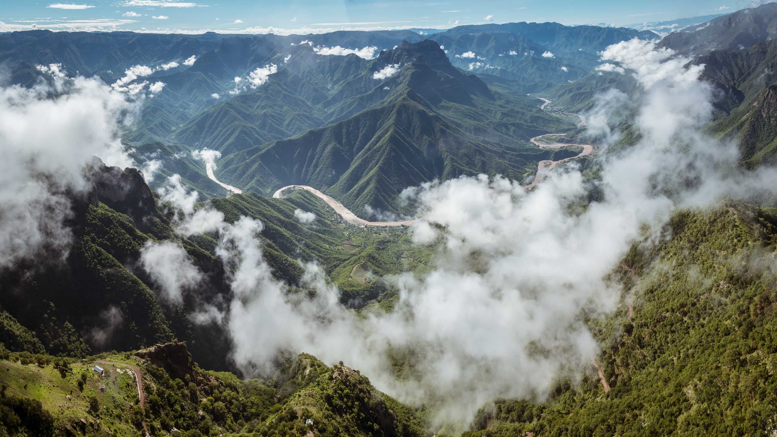 Vista aérea de las Barrancas del Cobre con nubes por debajo del mirador y el río serpenteando en el fondo del cañón.